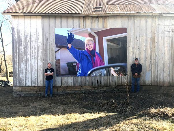 Two men stand near a barn with a large photo of a woman waving from a car.