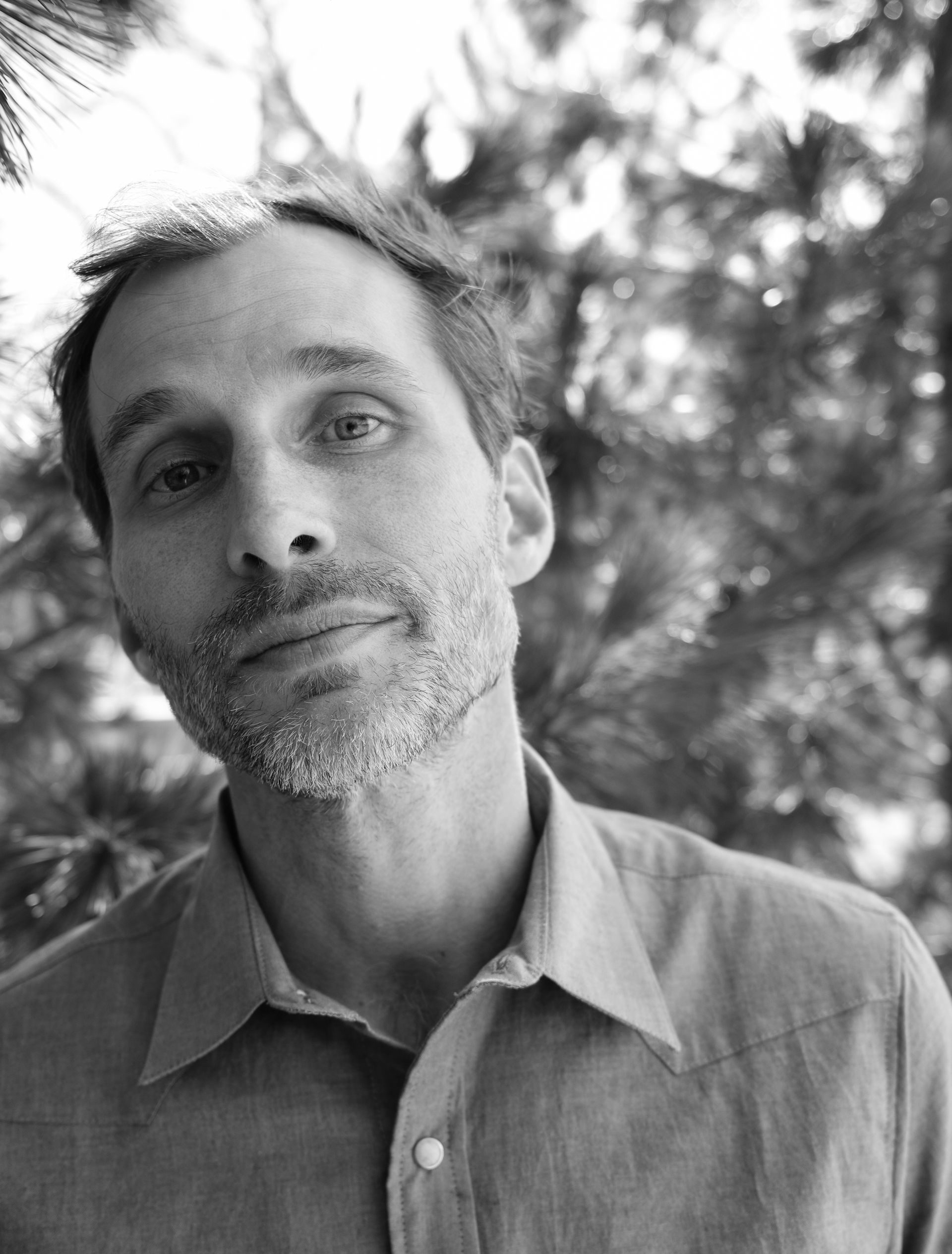 Man with a light beard and shirt looking towards the camera, against a blurry natural background.