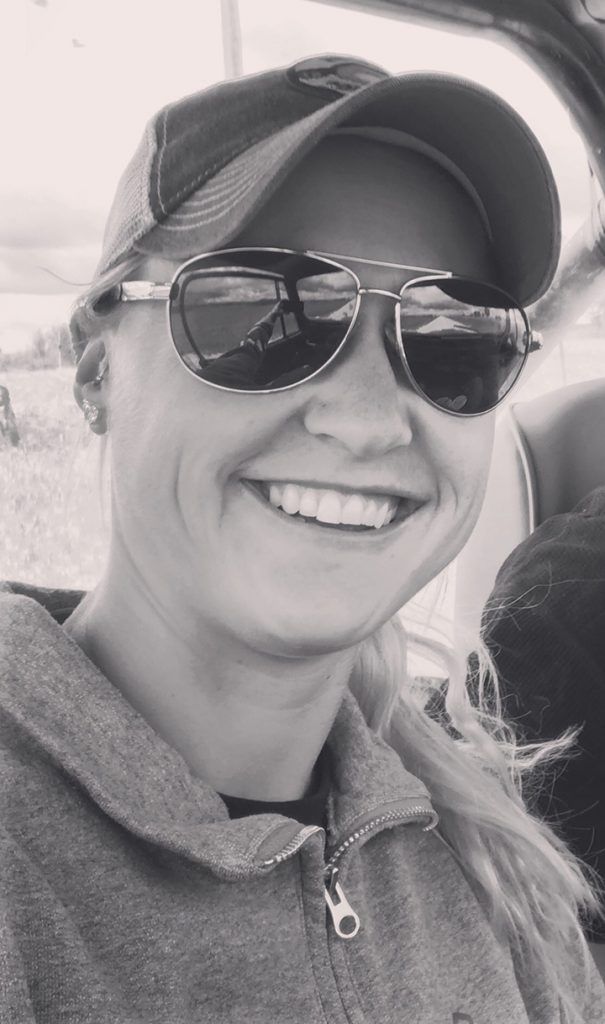 A woman wearing sunglasses and a hat is smiling in a black and white photo.