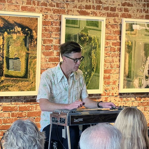 Man plays a pedal steel guitar at a venue with brick walls, with aerial photography art behind him.