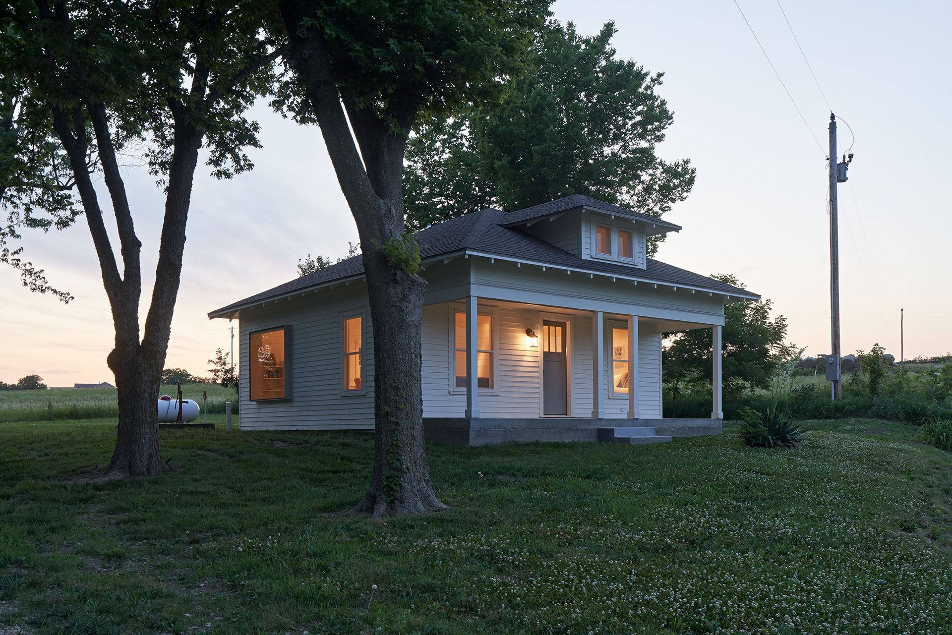 White cottage with porch and glowing windows, framed by trees in a grassy field. Dusk.