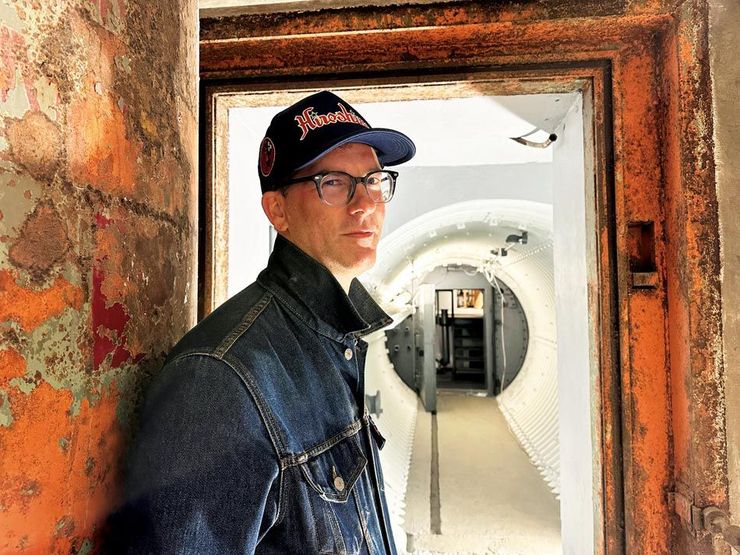Man in denim jacket and cap, standing in a doorway with peeling orange paint, looking toward a white-walled corridor.