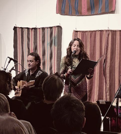 A woman sings into a microphone, holding a book, as a man plays guitar. They perform in front of a small audience.