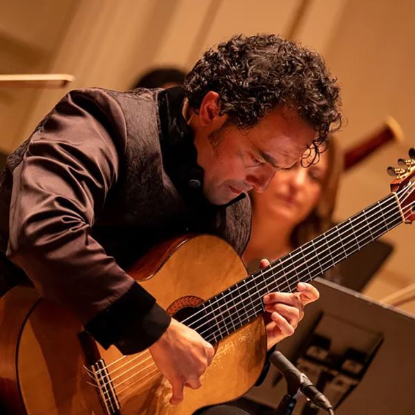 Man with curly hair playing a classical guitar, focused in an orchestra setting.