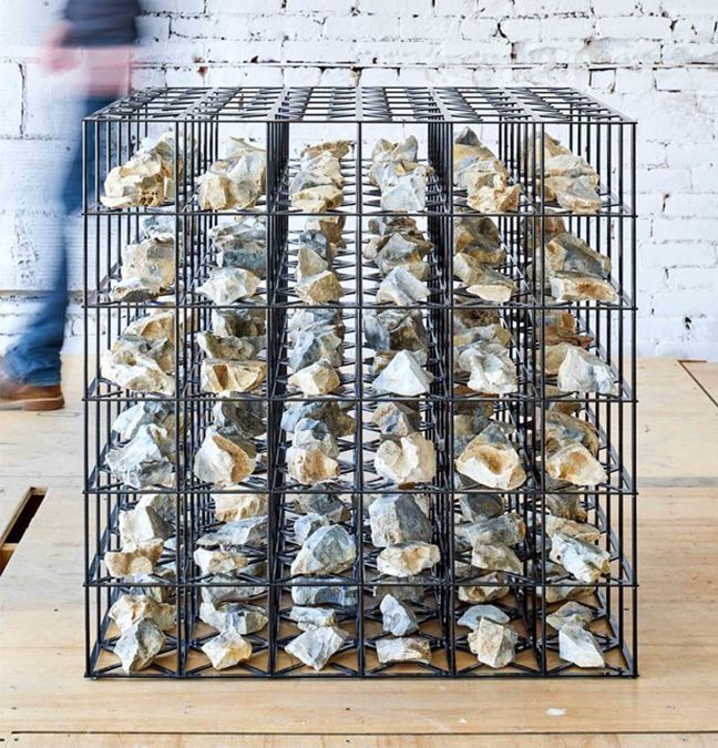 Black wireframe cube filled with gray rocks; person walks by in background.