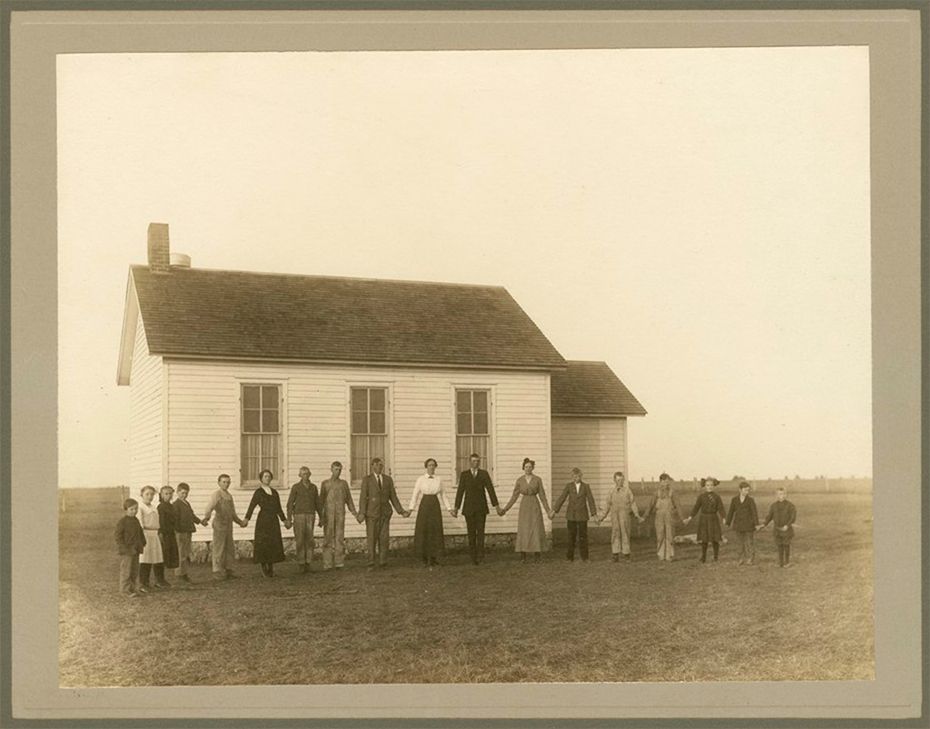 Group of people holding hands in front of a one-room schoolhouse, sepia-toned photograph.
