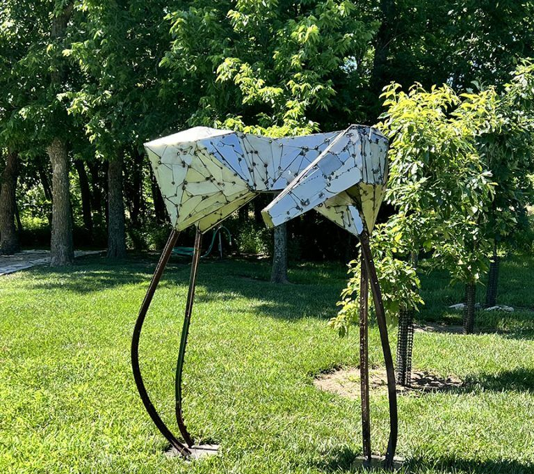 Metal sculpture of an abstract animal figure in a grassy park, legs are long and curved, head is geometric.