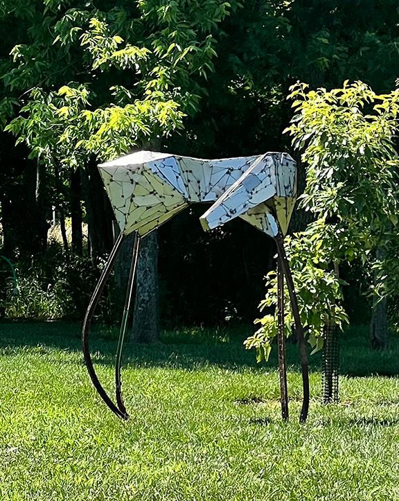 Geometric metal sculpture of an insect with long legs on a grassy lawn, trees in the background.