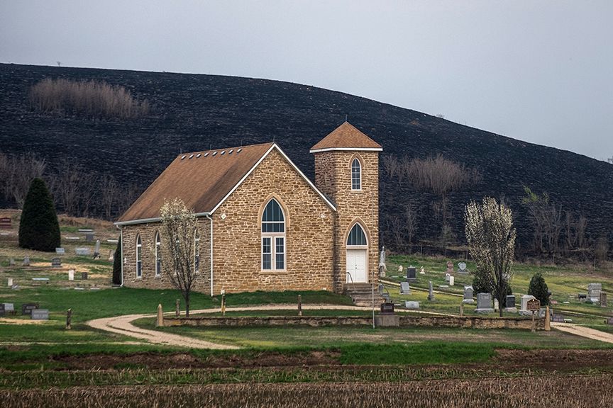 Stone church with a steeple and cemetery, set against a dark, charred hillside.