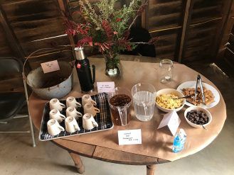 Coffee and breakfast spread on a table: cups, coffee, water, cereal, fruit, and a vase of flowers.