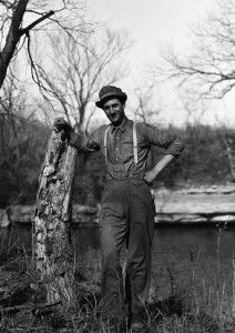 A black and white photo of a man leaning against a tree trunk.