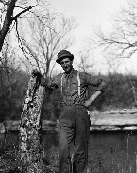A black and white photo of a man leaning against a tree trunk.