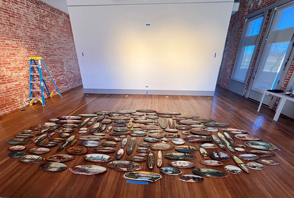 More than 100 log slab paintings arranged on the floor of the gallery in the shape of an oval, ready to be installed on the white wall of the gallery in the background.