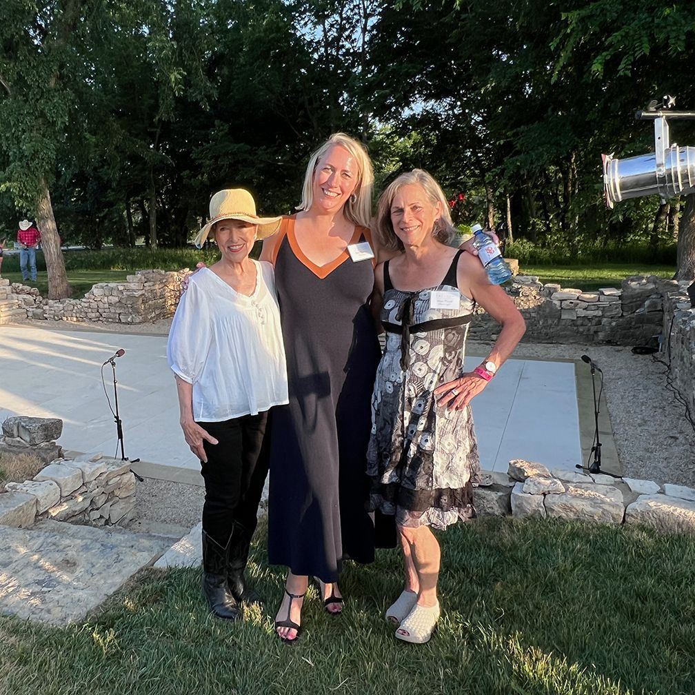 Three women posing in front of stone ruins. One wears a hat, one a long dress, and the third a floral dress.