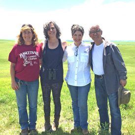 Four people stand in a field. Two women and two men pose for photo. Cloudy sky above green field.