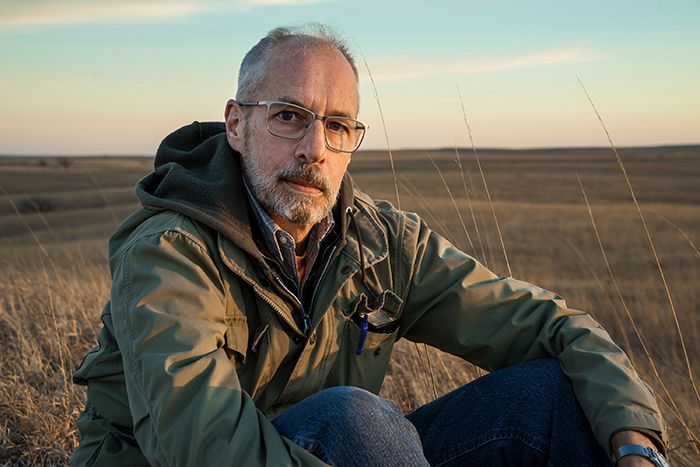 Man with glasses sits in tall grass, wearing a green jacket, looking at the camera, with a field backdrop.