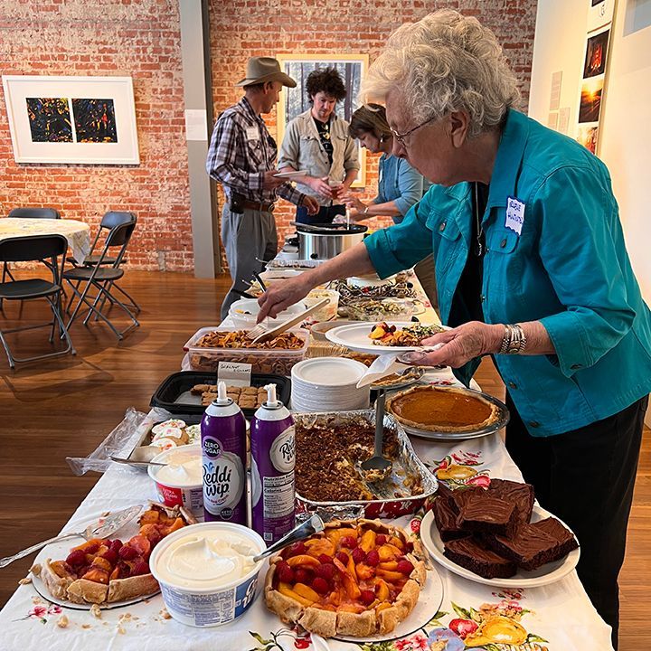 People serving themselves from a buffet table with pies, brownies, and fruit tarts in an art gallery.