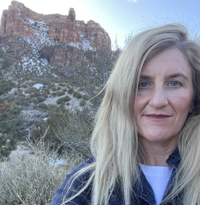Woman with blonde hair smiles in front of a snow-dusted red rock mountain.