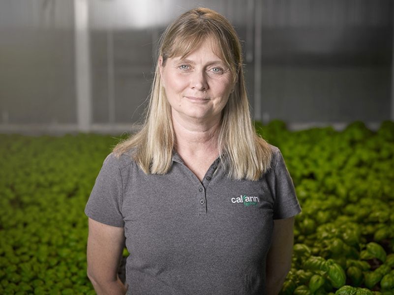 A woman in a gray shirt is standing in front of a field of green plants.