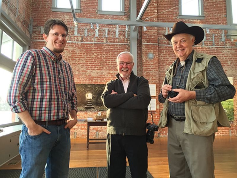 Three men standing indoors: one in plaid, one in a jacket, and one in a cowboy hat, smiling.
