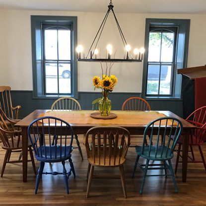 Dining room with a wooden table, colorful chairs, sunflowers, and windows.