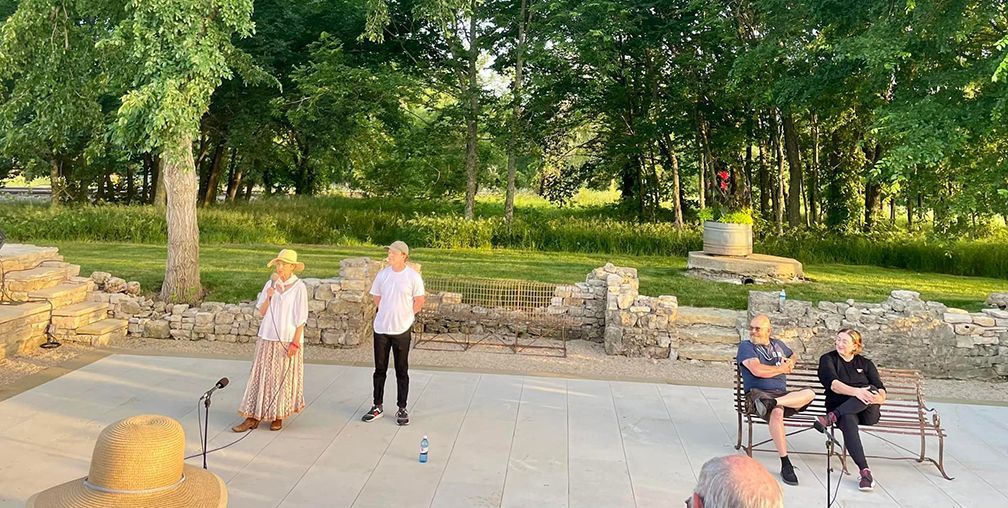 People standing and sitting in an outdoor area with stone ruins and trees.