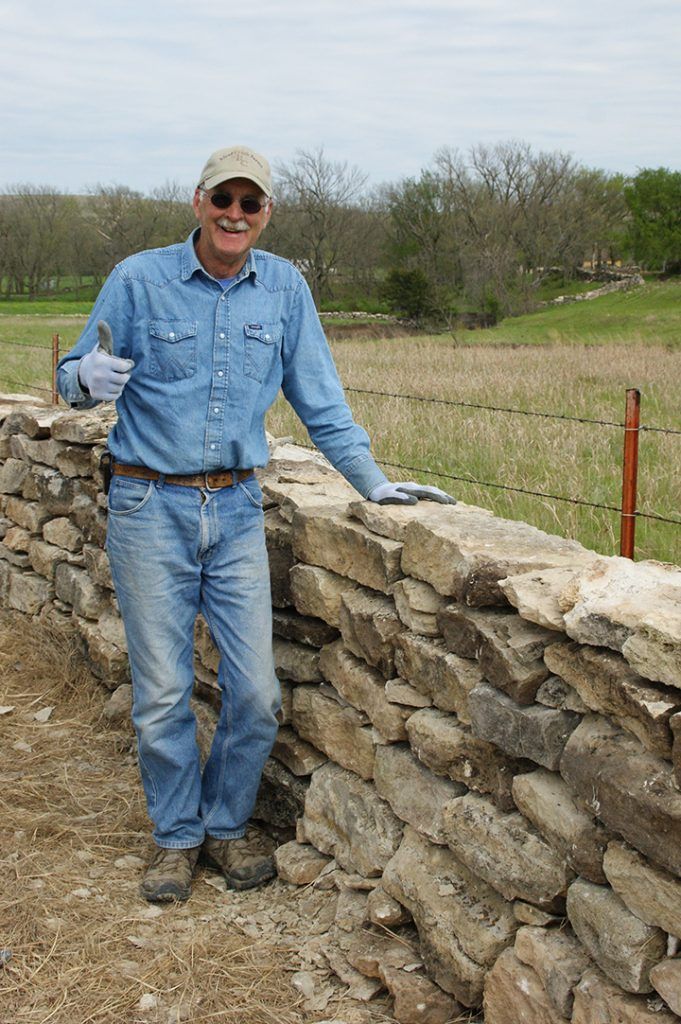 A man is standing next to a stone wall in a field.