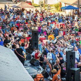 Crowd at outdoor concert; stage with band, people watching, raising arms, sunny day.
