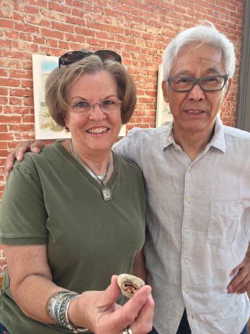 Woman holding small object with man beside her, brick wall background. Both are smiling, outdoors.