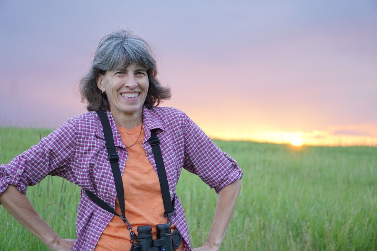 Woman with gray hair smiles outdoors, wearing binoculars. Sunset in background.