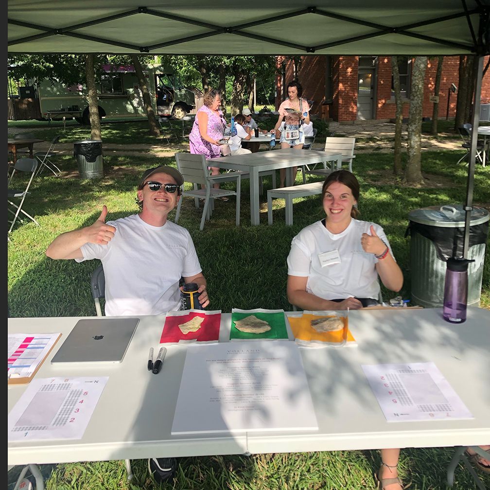 Two people at a table outdoors, smiling and giving thumbs up. Others sit at a table in the background.