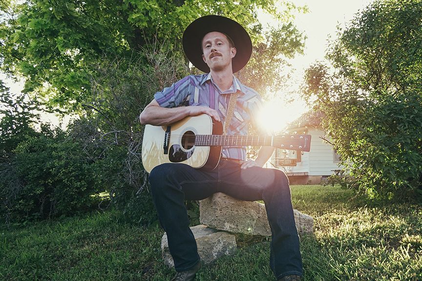 Man with guitar sits outside in sunlight, wearing hat and blue shirt.
