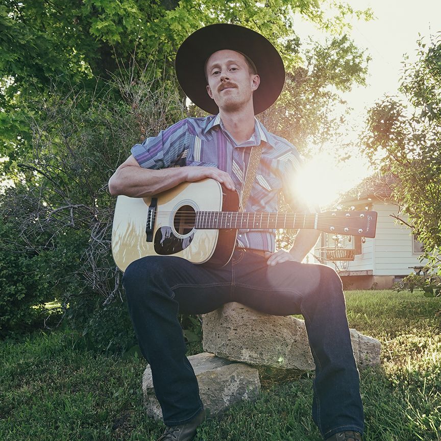 Man in cowboy hat plays acoustic guitar outdoors in sunlight.