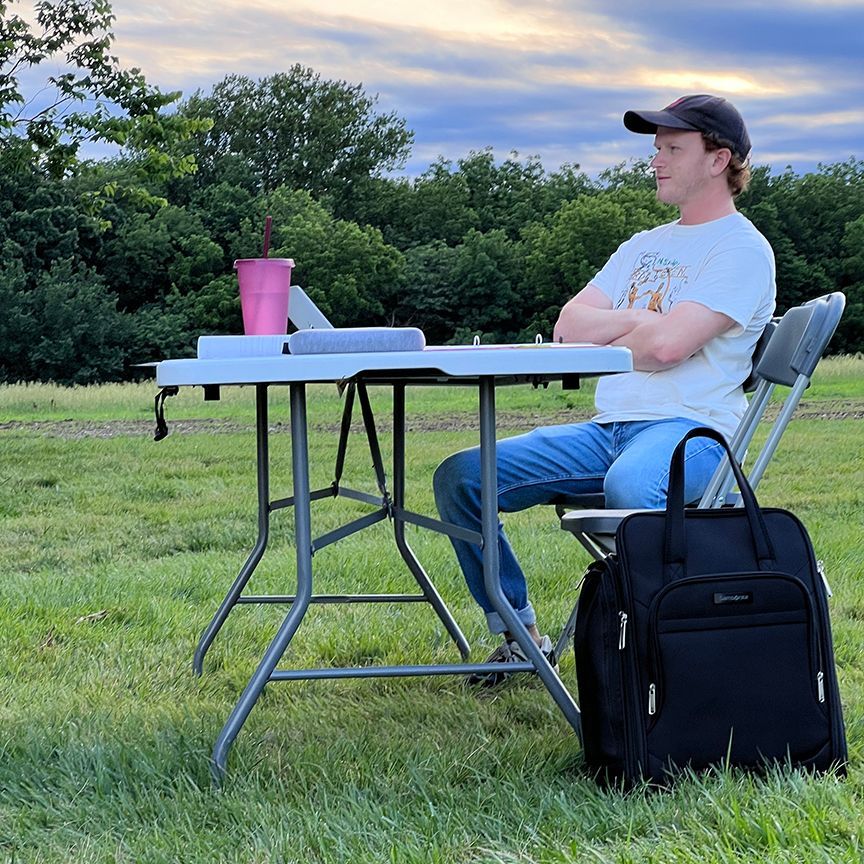 Man sits outdoors at a folding table, laptop and drink beside him. He wears a baseball cap and jeans.