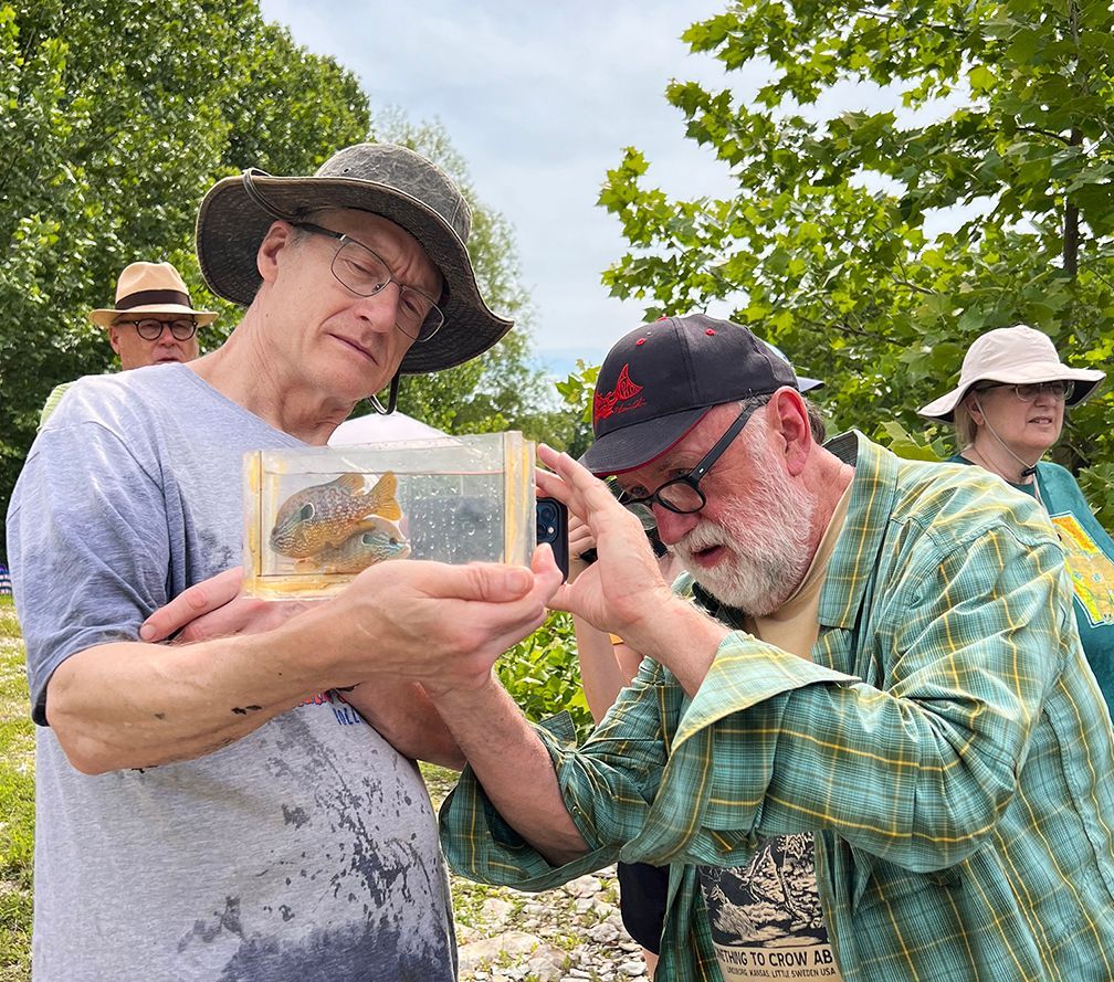 Two men examining an object in a small box, outdoors. One wears a hat, the other has a beard; other people are visible in the background.