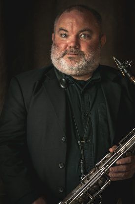 Man with a beard in black suit, holding a saxophone, against a dark background.