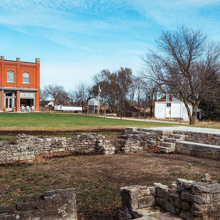 Ruins of stone foundation in foreground, red brick building, white building, green lawn, trees, blue sky.