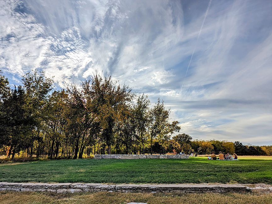 Lush green field with trees under a cloudy sky. Workers near a small building in the distance.