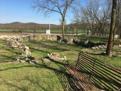 Stone ruins in a grassy area, a bench in the foreground, trees, and a distant hillside under a blue sky.