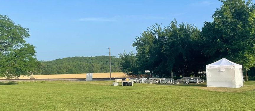 A field with a white tent, trees, and tables. Blue sky in background.