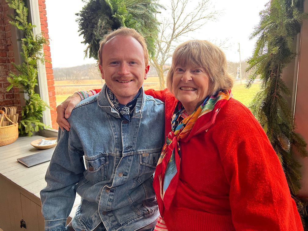 A man and woman seated on a window seat in front a a big window, with a Christmas wreath in the window and the landscape of the lawn beyond. 