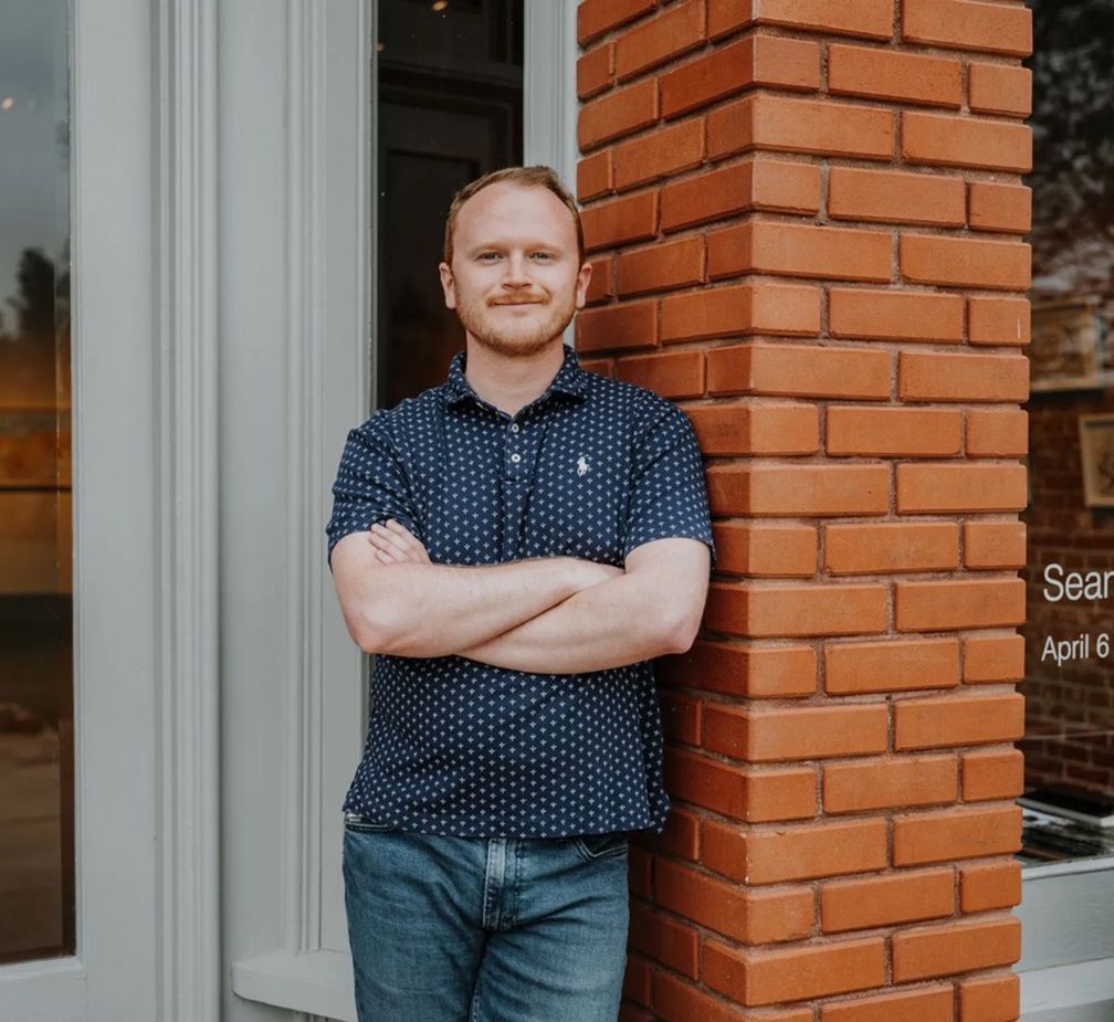 Man with arms crossed, leans against brick column by a white door. He wears a blue shirt and jeans.