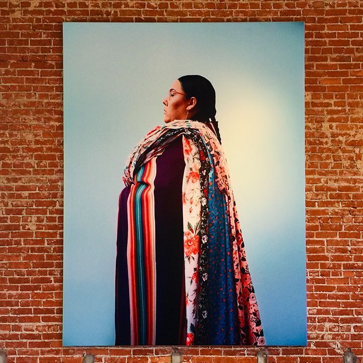 Woman in patterned shawl, standing against a light blue backdrop, mounted on a brick wall.