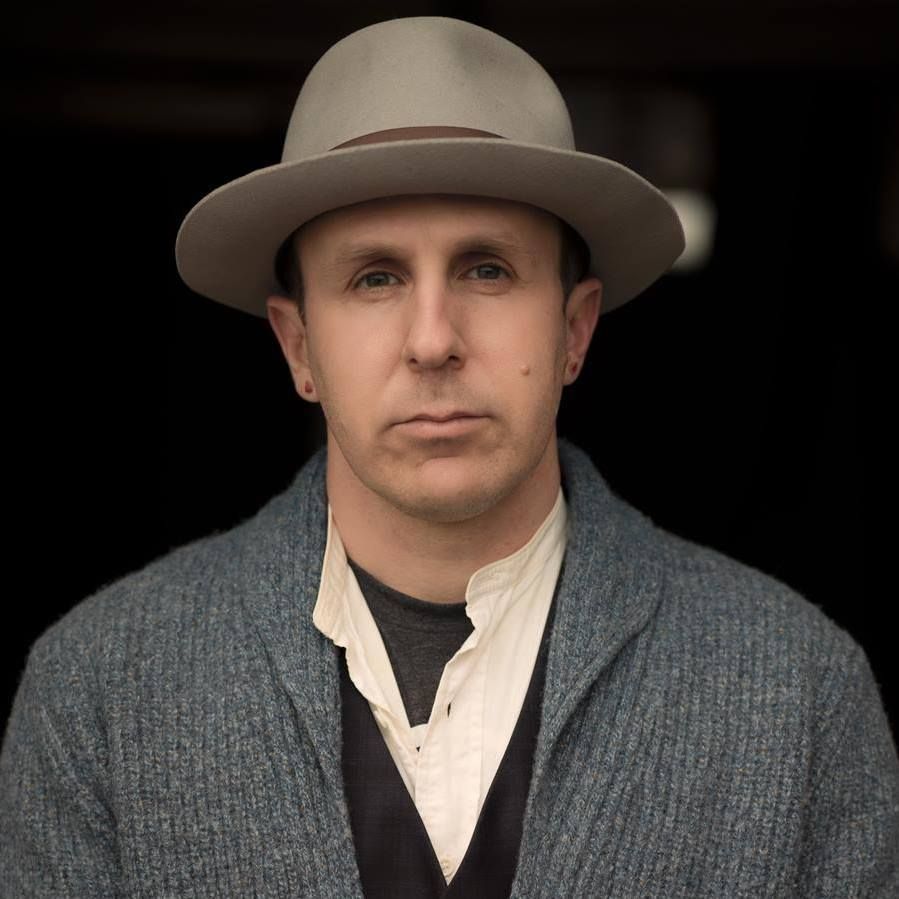Man wearing gray hat, blue cardigan, dark shirt, and white shirt, looking forward with neutral expression.