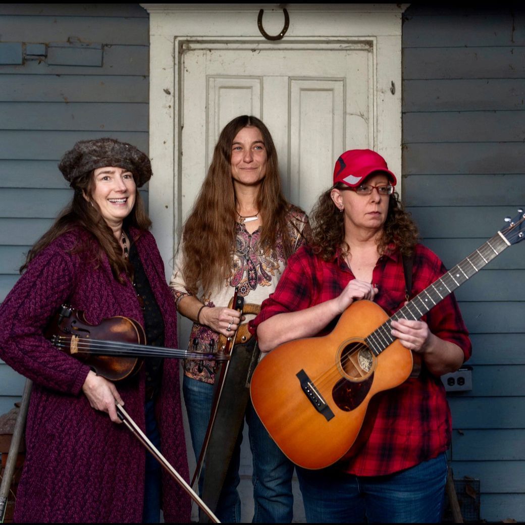 Three women, smiling, holding instruments, in front of a blue building: violin, banjo, and guitar.
