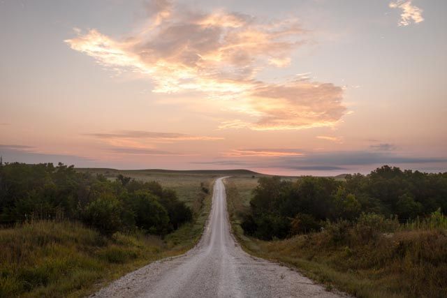 Gravel road disappearing into the horizon at sunset; pink and gold clouds above, trees on either side.