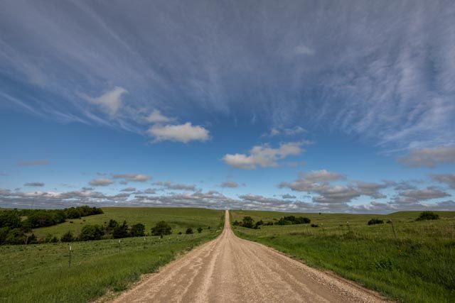Dirt road stretching to the horizon through a green field under a blue sky with streaky clouds.