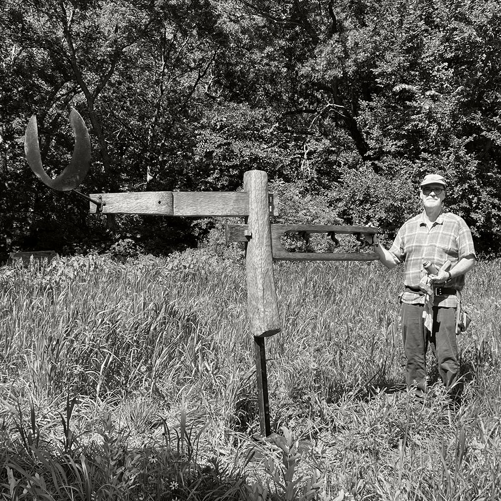 Man standing next to a rustic metal sculpture in a field, with trees in the background.