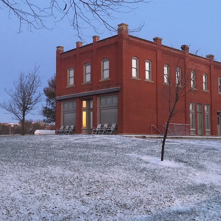 Red brick building with snow-covered lawn at dusk. Bare trees and chairs are visible.