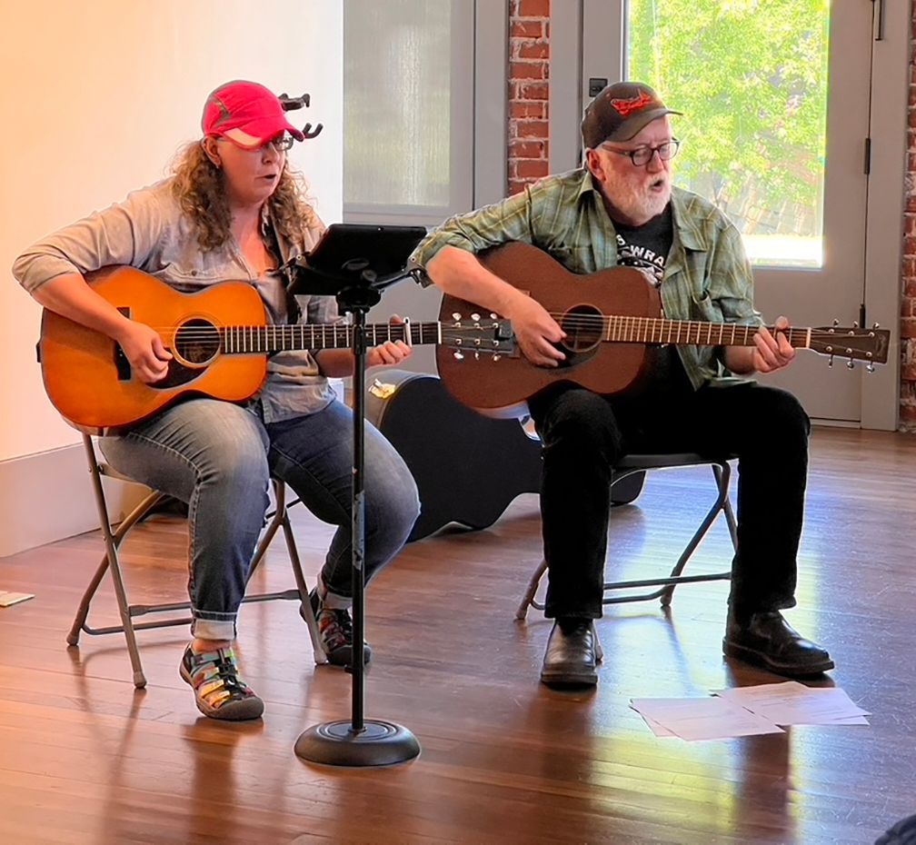 Two people, a man and a woman, playing guitars and singing while seated indoors.
